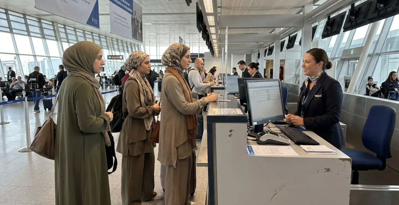 Deux femmes musulmanes en hijab et tenues pudiques élégantes discutent de profil à un comptoir d'aéroport moderne, baignées de lumière naturelle