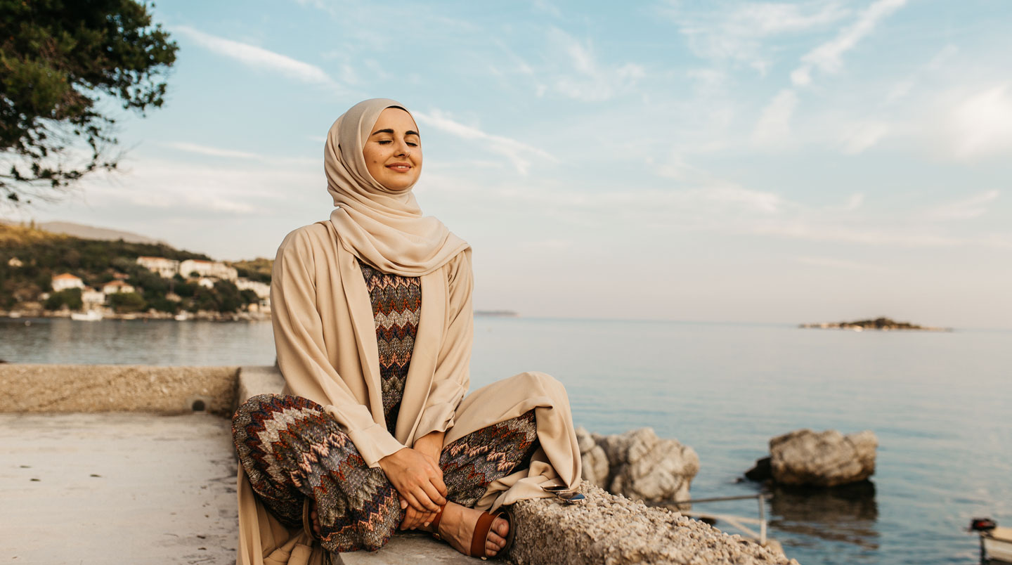 Femme en abaya beige vue de dos contemplant un paysage urbain ensoleillé depuis une fenêtre